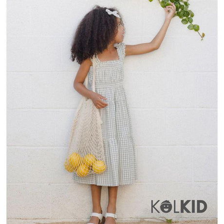 Young girl in a gingham sundress with white sandals and a headband holds a netted bag filled with yellow lemons while reaching for green leaves against a white wall; Kolkid logo in corner.