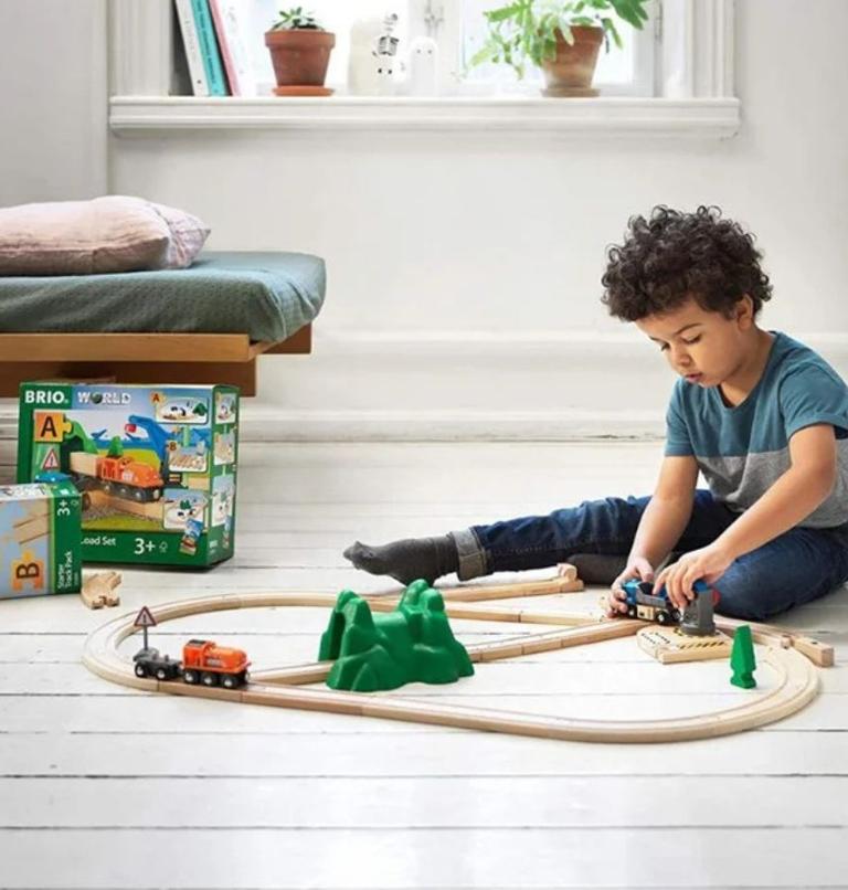 Young child sitting on a wooden floor playing with a wooden train track set and toy trains from Brio World in a bright indoor room