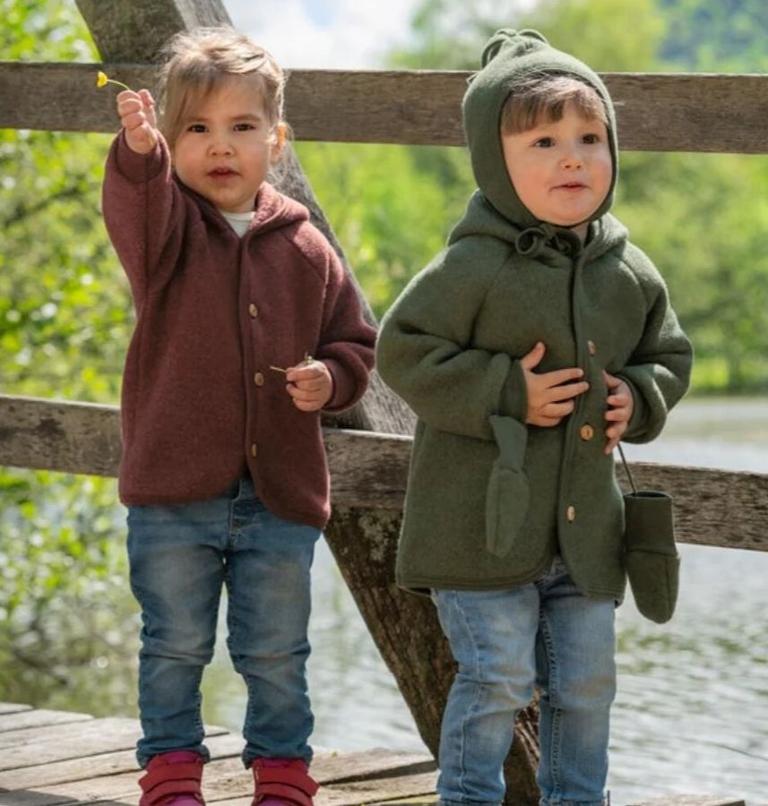 Two toddlers wearing Engel hooded wool jackets with wooden buttons from Kolkid, standing outdoors on a wooden bridge.