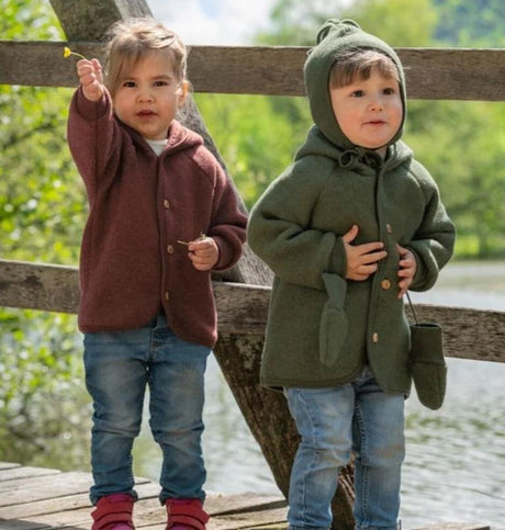 Two toddlers wearing Engel hooded wool jackets with wooden buttons from Kolkid, standing outdoors on a wooden bridge.