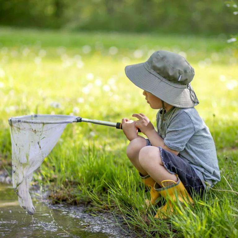 A young child crouches by a grassy pond, wearing a wide-brimmed olive sun hat, striped t-shirt, and yellow rubber boots, while holding a net to scoop water — showcasing outdoor exploration gear from Kolkid.