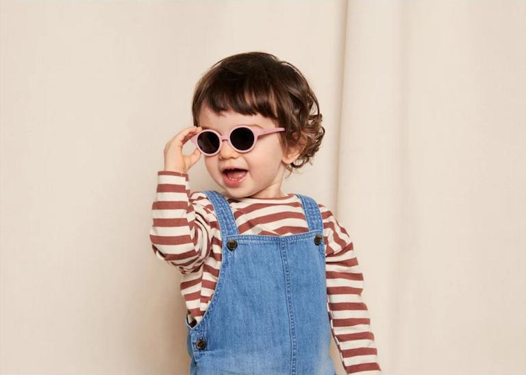 Smiling toddler wearing pink sunglasses, a striped shirt, and denim overalls, showcasing stylish and playful children's fashion from Kolkid.
