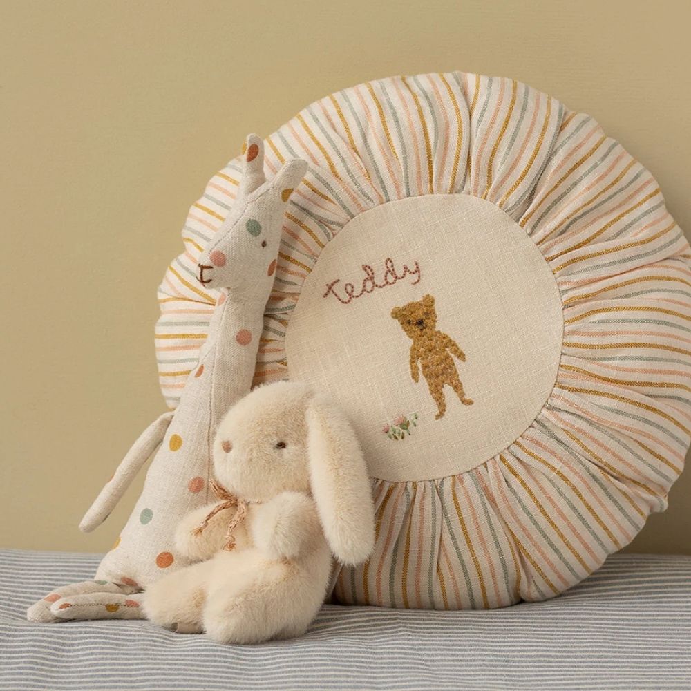 Round striped pillow with embroidered teddy bear design, accompanied by plush toys on a striped surface.