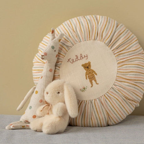 Round striped pillow with embroidered teddy bear design, accompanied by plush toys on a striped surface.